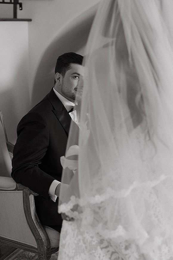 Ceremony moment as groom in black tuxedo and bow tie looks up from wooden chair at bride’s lace dress and long veil in indoor aisle