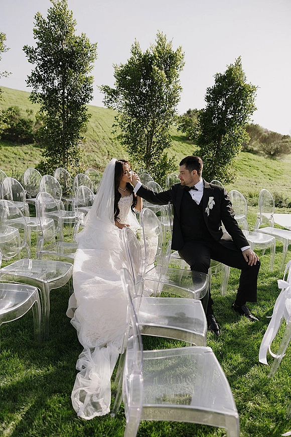 Couple portrait of bride and groom seated as he adjusts her hair, her veil draped over clear acrylic chairs on a grassy hillside lawn
