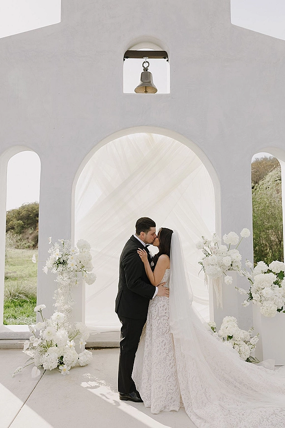 Wedding kiss as bride and groom embrace at the altar beneath a white arch with floral arrangements and a flowing veil, stucco wall behind