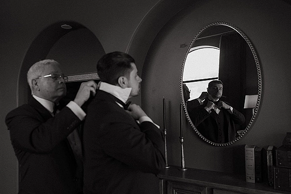 Groom getting ready, adjusting bow tie in a classic black tuxedo by a mirror, soft window light near an arched doorway and dresser
