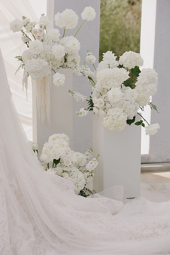 Wedding floral arrangement of white hydrangeas and roses on modern pedestal plinths, with soft draped fabric and window light