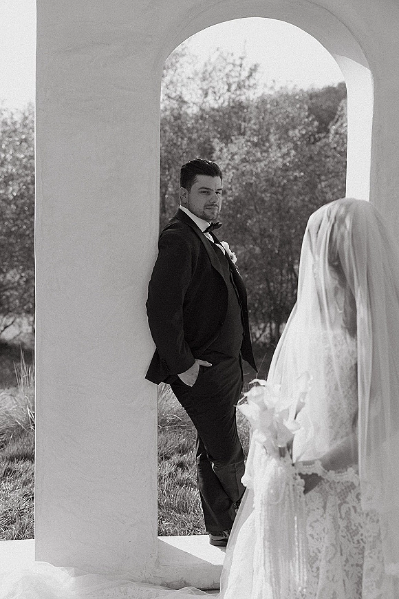 First look moment as bride in lace veil approaches groom in black tuxedo with bow tie, under a stone archway with trees behind