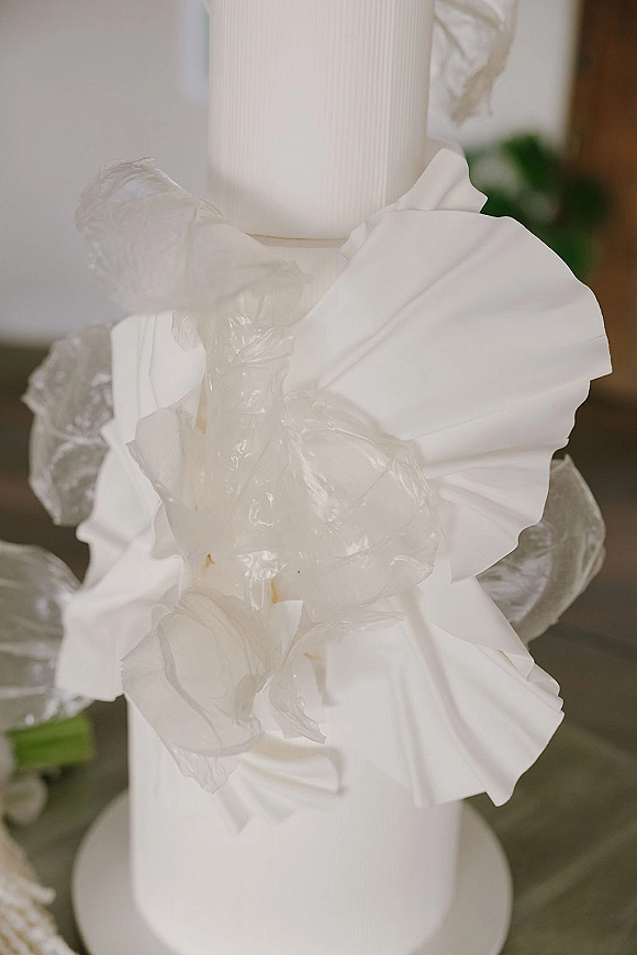 Wedding candle decor with a white pillar candle centerpiece wrapped in translucent floral fabric and ribbon on a tabletop with blurred greenery