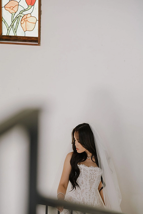 Bridal portrait of a bride in veil wearing a strapless lace wedding dress with pearls, leaning on a railing near stained glass indoors