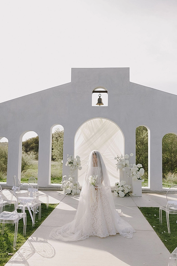 Bridal portrait of a bride holding bouquet in a cathedral veil, standing before a white chapel arch with draped altar and florals