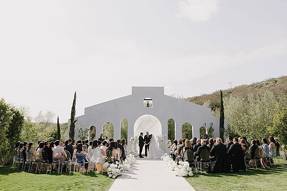 Wedding ceremony setup with a white aisle runner lined by floral arrangements, clear chairs for guests, and a white arch on a hillside lawn