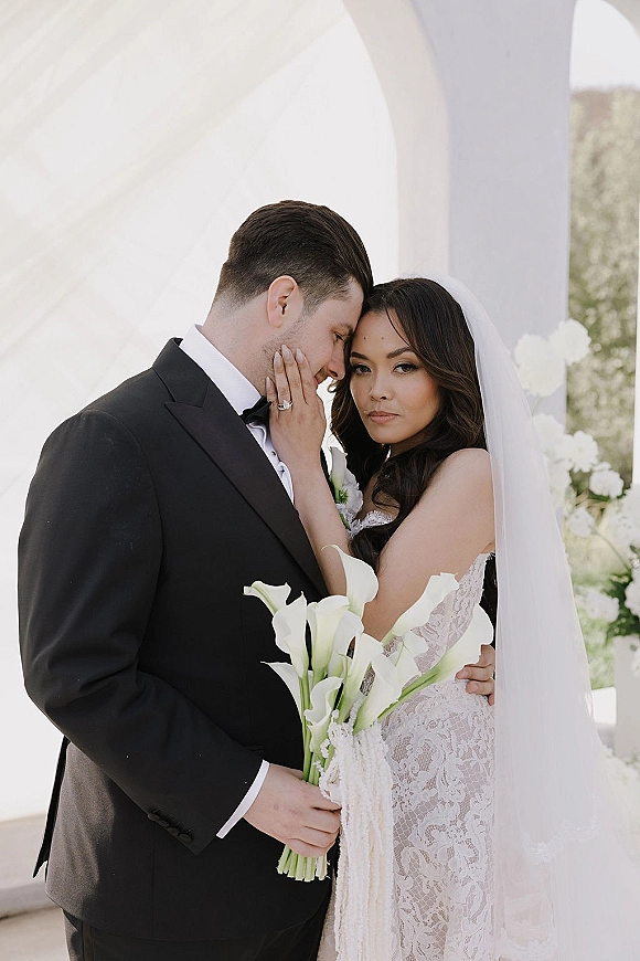 Couple portrait of bride and groom embrace under a white-draped wedding arch, her veil and lace dress framing a calla lily bouquet