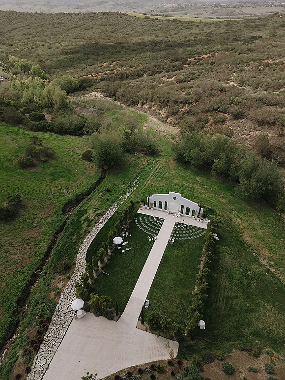 Outdoor ceremony setup with a white wedding arch and aisle runner, curved chairs and florals on a stone walkway in a hillside field