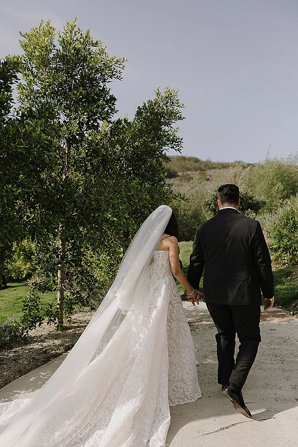 Couple portrait of newlyweds holding hands walking away, bride’s long veil trailing behind on a tree-lined garden path