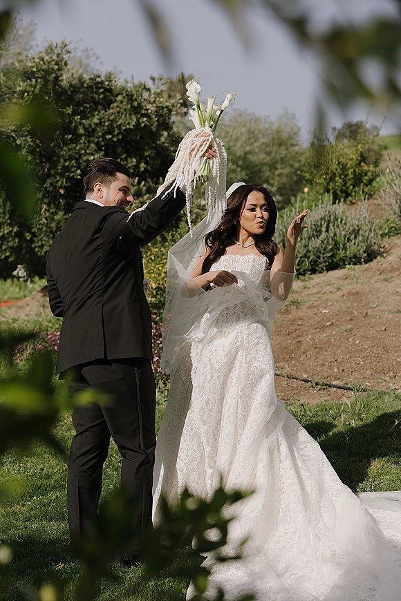 Wedding couple portrait with bride blowing a kiss as groom lifts a calla lily bouquet overhead in a sunny garden with green shrubs