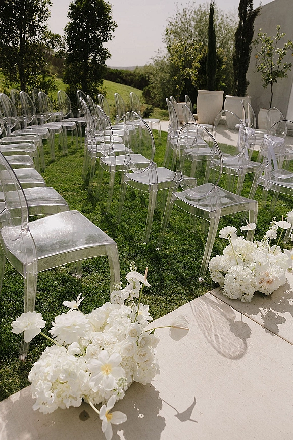 Ceremony seating with clear acrylic wedding chairs tied with white ribbon, flanked by white florals on a grassy lawn with hills