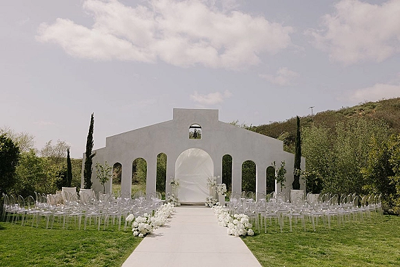 Ceremony setup for an outdoor wedding ceremony with white aisle runner and clear acrylic chairs facing an arched backdrop on a green hillside lawn