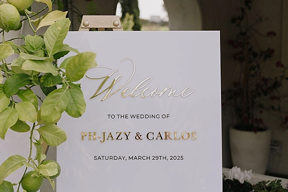 Wedding welcome sign with calligraphy and gold lettering on an easel, framed by greenery leaves near an outdoor doorway entrance