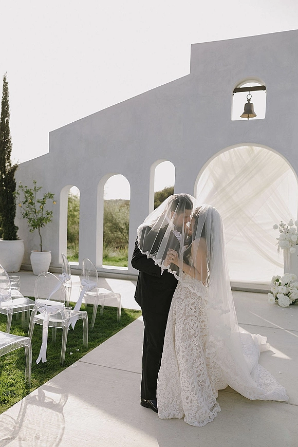 Wedding kiss as bride and groom kissing under a draped arch, cathedral veil trailing over lace gown on a white stucco courtyard lawn