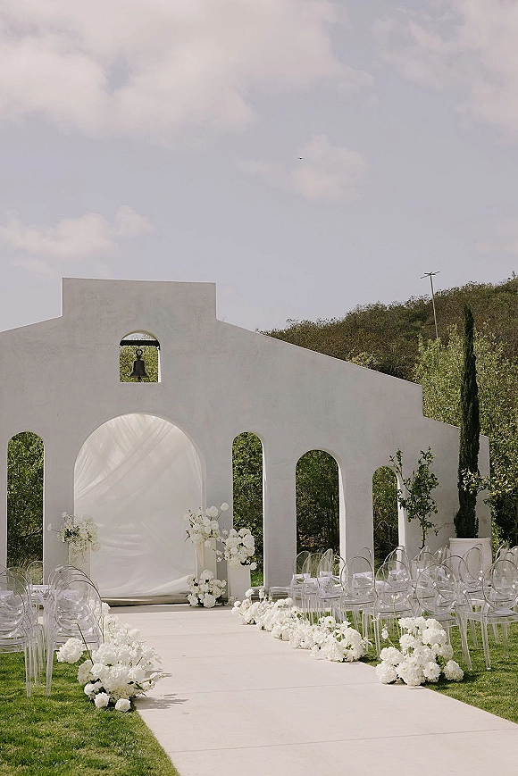 Ceremony setup for outdoor wedding ceremony with a white wedding aisle runner, clear acrylic chairs, and floral arch at a stucco chapel façade