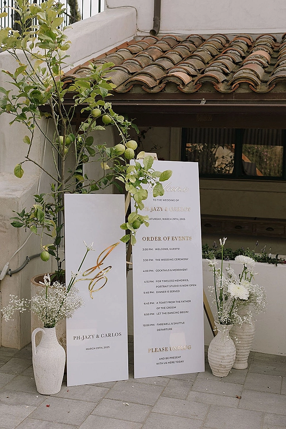 Wedding signage display with a wedding welcome sign and timeline boards, styled with white vases, baby’s breath, greenery, and a lemon tree on a stucco patio