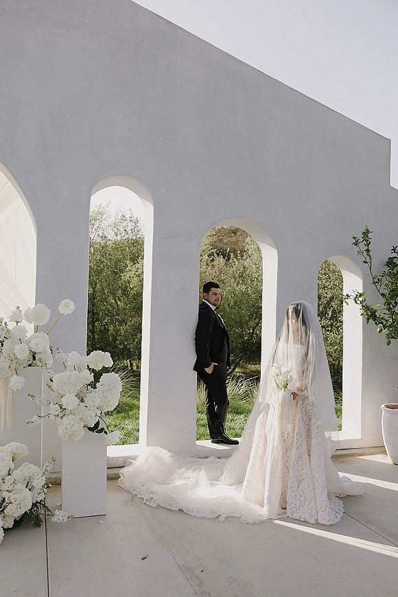 Wedding first look as groom in a black tux waits while bride in a lace dress and veil approaches with a white bouquet beneath stucco arches