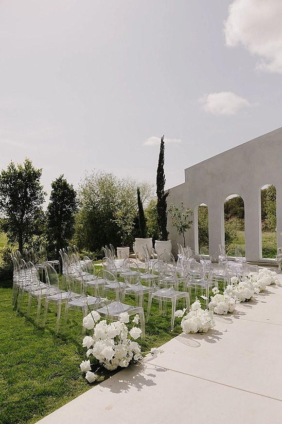 Ceremony setup for an outdoor wedding ceremony with clear acrylic chairs and white aisle flowers on a lawn beside a white stucco arched wall