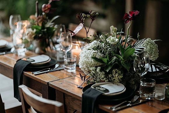 Reception tablescape on a rustic wedding table with white hydrangeas and greenery centerpiece, taper candles, and black place settings in dim light