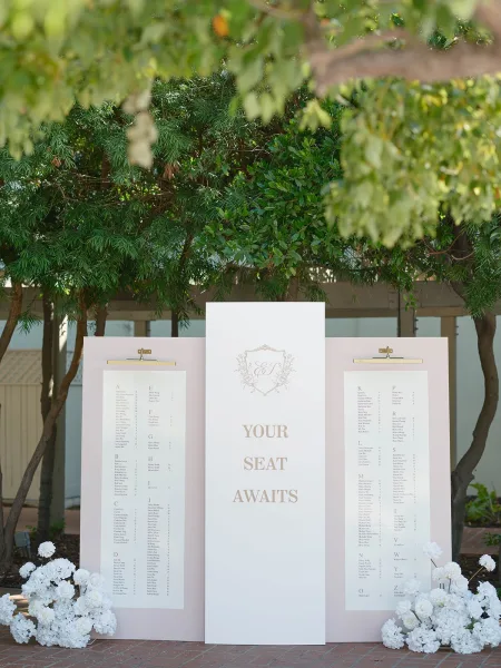 Wedding seating chart displayed on foam boards with clip-on sign lights and white floral accents along a brick walkway by greenery
