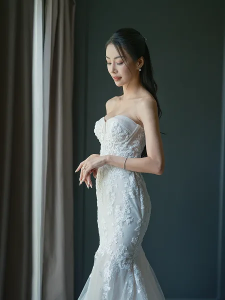 Bridal portrait of a bride by window in a strapless lace mermaid gown with drop earrings, shown in soft window light by curtains and a dark wall