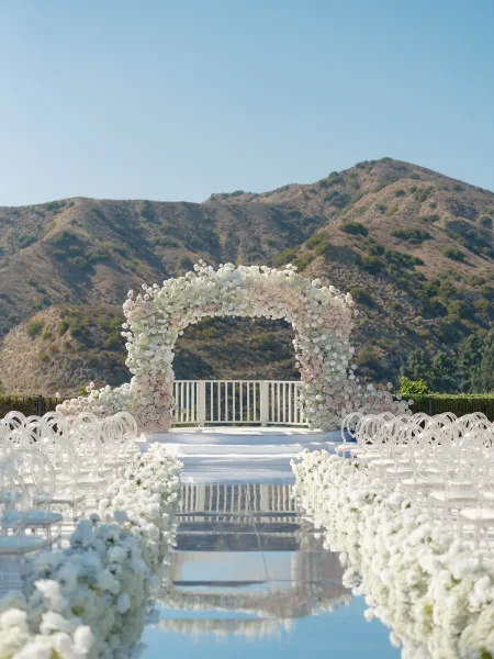 Ceremony setup with an outdoor wedding ceremony setup featuring a floral arch, blush and white aisle florals, and clear chairs on a mountain terrace