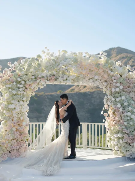 Wedding kiss under a round floral arch of white and blush roses, bride in lace gown and veil with mountain landscape beyond the terrace