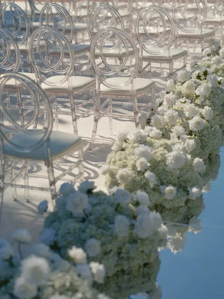Ceremony aisle decor with clear acrylic wedding chairs, a white aisle runner, and lush white roses and hydrangeas lining outdoor seating rows