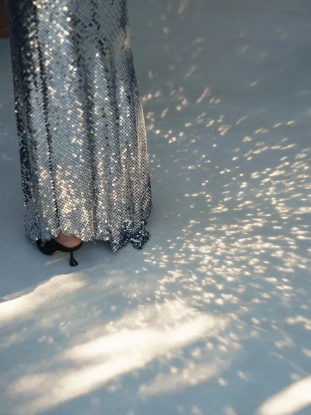 Sequin dress detail of a silver sequin wedding dress hem with black kitten heels on sunlit pavement in dappled tree shadows