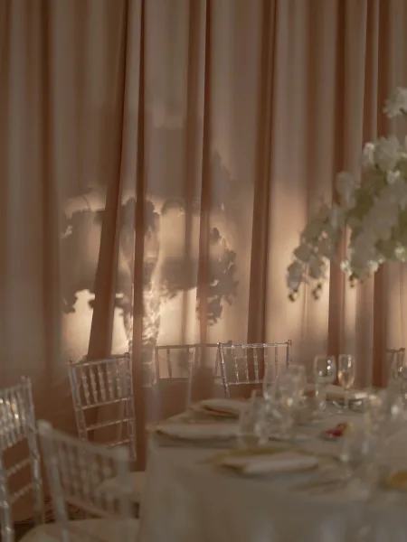 Reception tablescape with round wedding table setting, white linens, champagne flutes and floral centerpiece beneath draped backdrop and warm uplighting