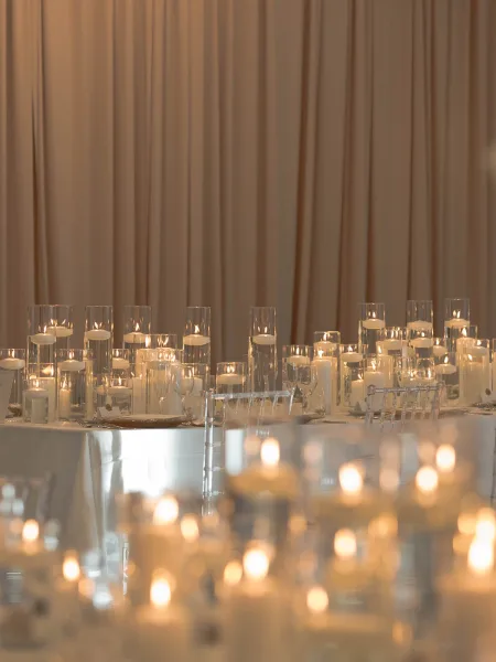 Reception tablescape with floating candle centerpiece in glass cylinder vases, white linens, and clear chiavari chairs before a draped backdrop