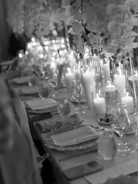 Reception tablescape with pillar candle centerpiece, white floral centerpieces, glass vases, and place settings on a long banquet table in dim light