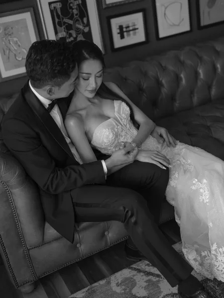 Couple portrait in a black and white wedding portrait, groom kissing bride’s cheek as they hold hands on a tufted leather sofa indoors