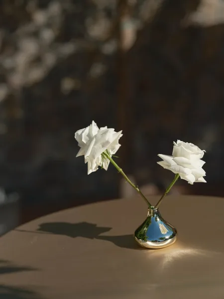 Floral centerpiece with a single white rose in a metallic bud vase on a tabletop, set against soft shadows and a dark blurred backdrop