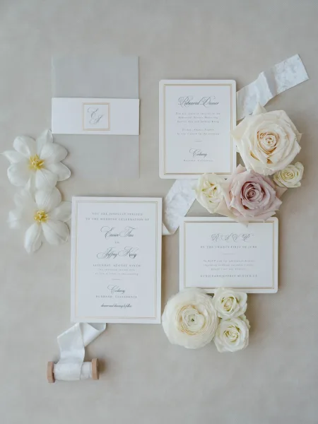 Invitation suite flatlay with wedding invitation set, monogram and RSVP cards tied in vellum ribbon with roses and ranunculus on linen backdrop