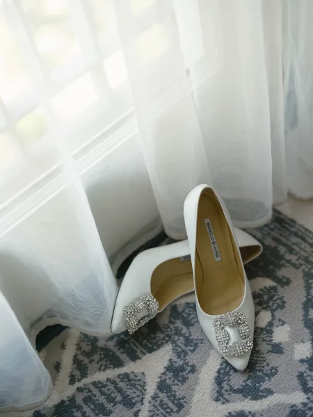 Bridal shoes, white wedding heels with crystal buckle clips resting on a patterned rug by a window, softly lit through sheer curtains