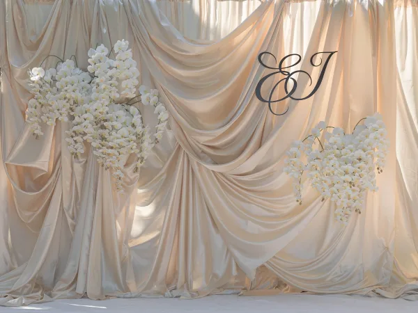 Wedding backdrop with draped wedding backdrop fabric, white orchid accents and a monogram sign set against sheer curtains and a white floor