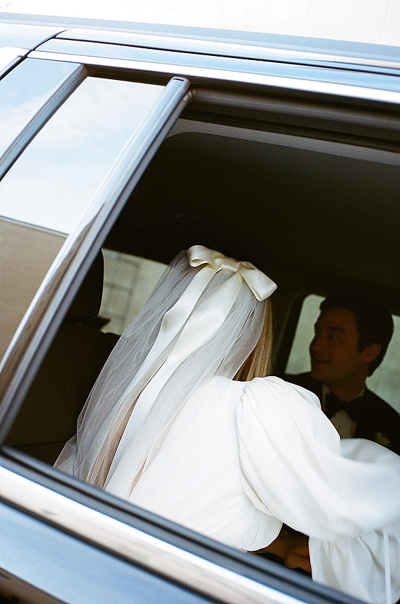 Wedding getaway car moment with bride in car wearing a cathedral veil and oversized hair bow beside groom in tux, framed by the window