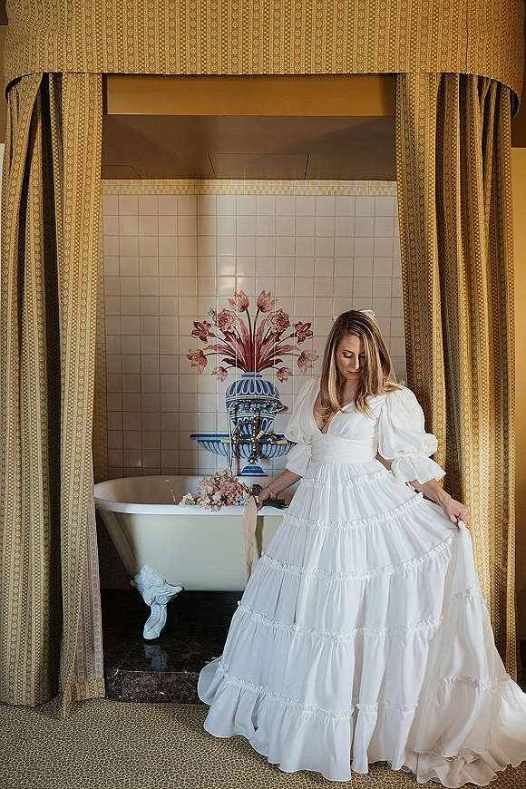Bridal portrait of a bride in a wedding dress with puff sleeves holding a bouquet with ribbon beside a clawfoot tub in a tiled bathroom alcove