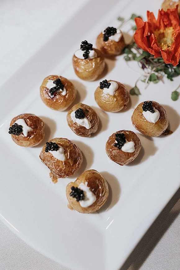 Wedding hors d'oeuvres of potato and caviar bite topped with sour cream on a white plate, accented with an orange flower on linen