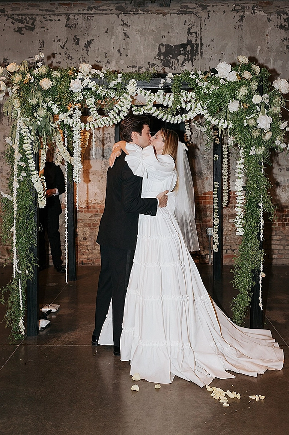 Wedding kiss portrait of bride and groom kissing under a floral arch with hanging white flowers and rose petals in an industrial brick venue