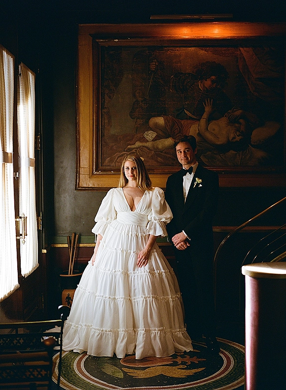 Couple portrait of bride and groom in formal attire, her puff sleeve gown and his tuxedo, posed by framed painting and staircase banister indoors