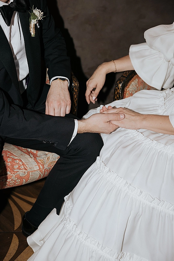 Couple holding hands, wedding hand holding photo showing bride’s dress and groom’s black tuxedo with boutonniere, seated indoors on a vintage chair