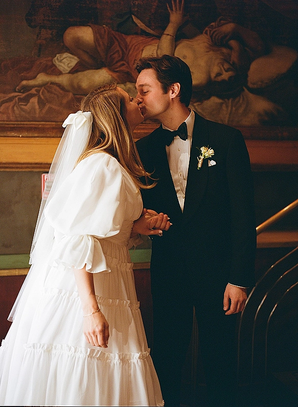 Wedding kiss portrait of bride and groom kissing, her bow veil and white gown beside a stair railing with a mural backdrop