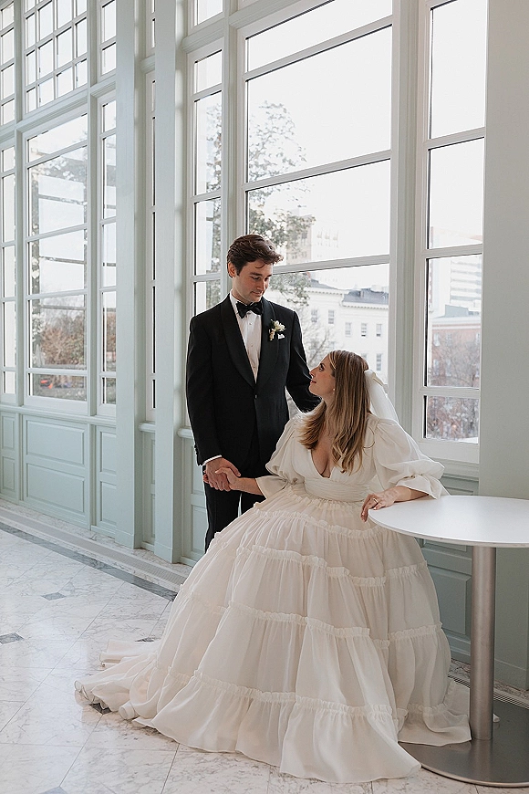 Couple portrait with bride and groom holding hands, bride in veil looking up at tuxedo groom in a windowed marble hallway.
