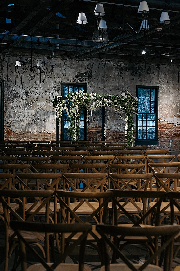 Ceremony setup with a floral arch and greenery garland, white blooms and cross back chairs before a brick and plaster wall with chandeliers