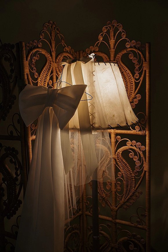 Wedding dress detail with bridal veil detail, featuring a white bow and tulle veil on a hanger beside a floor lamp against a dark wall
