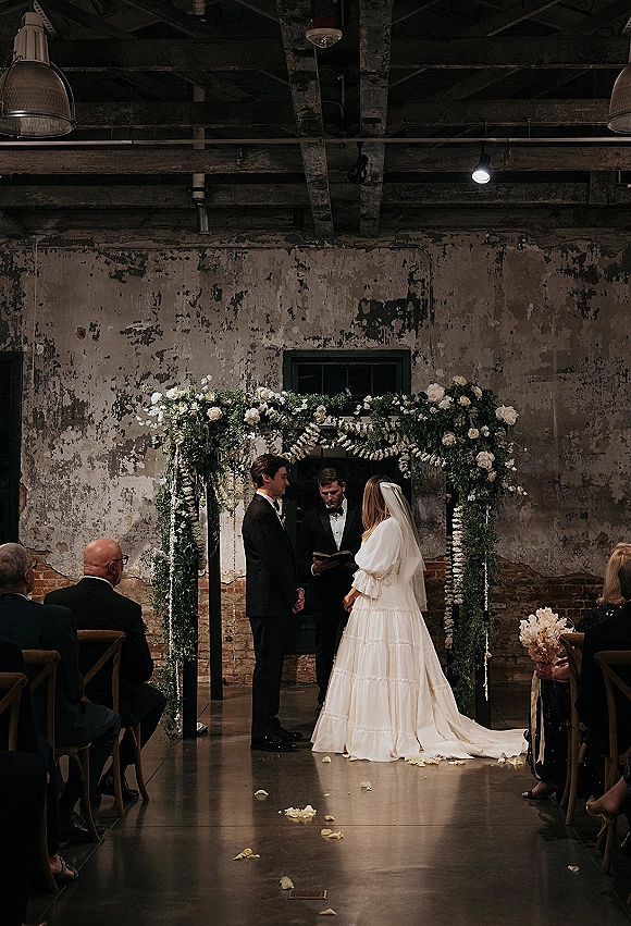 Wedding ceremony with couple exchanging vows beneath a white flower arch and hanging florals, rose-petal aisle in an industrial brick venue