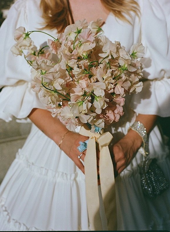 Bridal bouquet of sweet peas with blue accent flowers and long cream ribbon wrap, held against a white dress indoors by a neutral wall