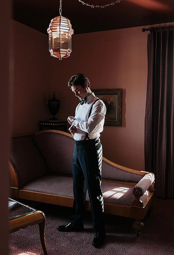 Groom portrait of a man adjusting cufflinks in a white shirt with bow tie and suspenders, posed by a vintage sofa under lantern light